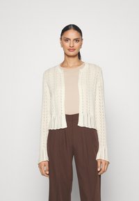 Knit cardigan in cream with an open-front design, featuring a textured pattern and ribbed cuffs, paired with a light beige top and brown trousers.