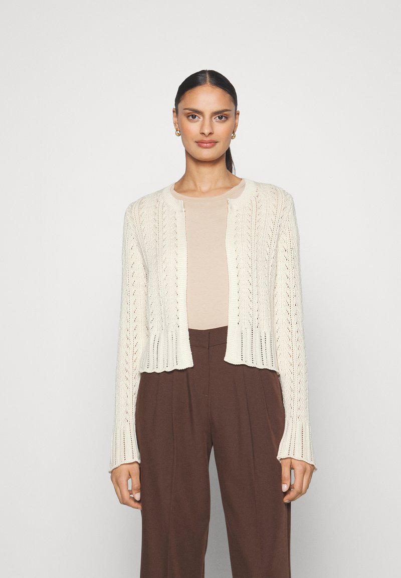Knit cardigan in cream with an open-front design, featuring a textured pattern and ribbed cuffs, paired with a light beige top and brown trousers.