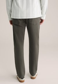 Man in een donkerolijfgroene straight-leg broek, een wit shirt met verticale strepen en witte sneakers met een bruine zool die staat.