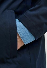 Marineblå udendørs jakke med en sidelomme, der afslører en lys blå denimskjorteærme. Stoffet ser glat ud med et struktureret design.