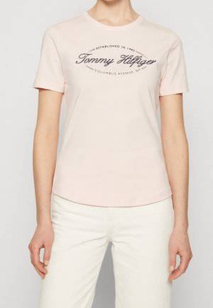 Kobieta ubrana w jasnoróżową koszulkę z krótkim rękawem z napisem „Tommy Hilfiger” oraz białe spodnie, stoi z ramionami rozluźnionymi po bokach.