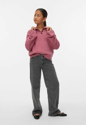 Joven con un suéter rosa de botones, jeans grises de pierna ancha y bailarinas negras de pie contra un fondo blanco liso.