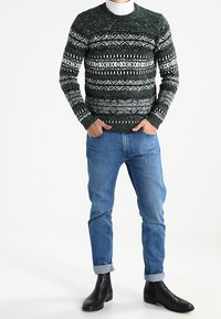 Jersey de punto con patrón verde y blanco y cuello redondo, combinado con vaqueros azules con dobladillo remangado y botines negros.