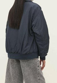 Marineblauwe, lichtgewicht bomberjack met elastische manchetten en zoom. Het heeft een soepele textuur en een relaxte pasvorm, gecombineerd met grijze wijde jeans.