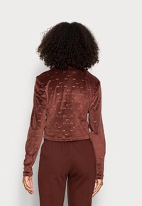 Kvinna med lockigt hår som bär en brun långärmad Nike-jacka och matchande byxor, sedd bakifrån mot en enfärgad bakgrund.