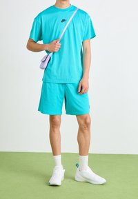 Turkos nättröja och shorts, med en liten lila axelremsväska. Vita skor med detaljer, stående på grön konstgräs.