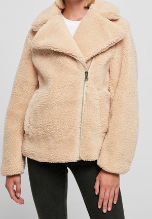 Femme portant une veste beige en fausse peau de mouton zippée avec un large col et un pantalon noir, debout devant un fond uni.