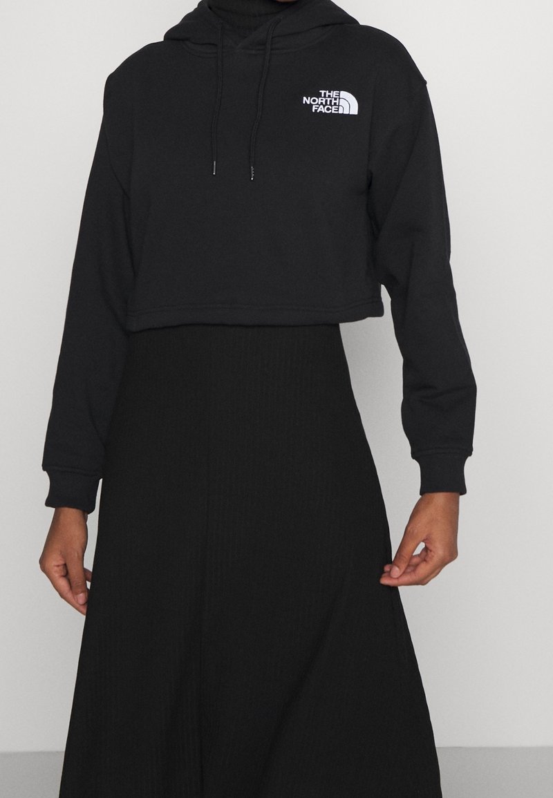 Černá zkrácená mikina s kapucí z měkké látky, vybavená přední kapsou typu klokanka, stahovací kapucí a bílým logem na levé straně hrudi.