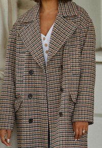 Dubbele rij knopen jas gemaakt van stof met een pied-de-poule patroon in bruin, crème en zwart. Voorzien van een grote kraag en twee voorzakken.