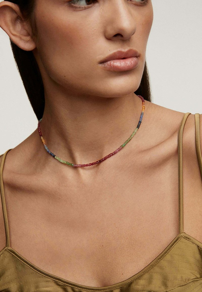 Collar de cuentas coloridas que presenta pequeñas cuentas redondas en varios colores, incluyendo rojo, verde, azul y amarillo, descansando sobre un cuello desnudo.