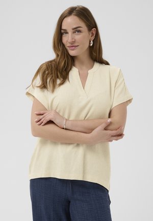 Žena so svetlohnedými vlasmi, oblečená v krémovom krátkokošele a tmavomodrých nohaviciach, stojaca so založenými rukami pred jednoduchým pozadím.