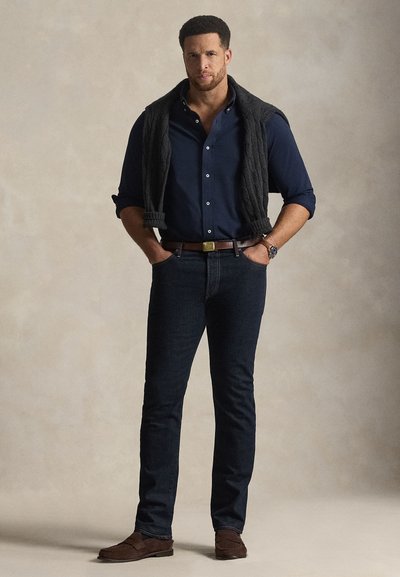 Muškarac s kratkom kovrčavom kosom i bradom stoji s rukama u džepovima, nosi mornarsko plavu košulju, tamne traperice, smeđi remen i cipele, s tamnim džemperom prebačenim preko ramena.