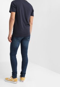 Camiseta de algodón azul marino, diseño ajustado, combinada con jeans ajustados de color azul oscuro y zapatillas amarillas con acentos y cordones blancos.