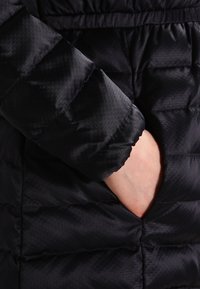 Svart pufferjacka med ärm som har ett texturerat tyg med hexagonala mönster och en sidoficka, vilket framhäver dess isolerade design.