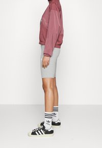 Lehká tmavě červená bunda, šedé přiléhavé šortky a černé tenisky s bílými detaily. Bílé ponožky s proužky a logem. Kompozice z bočního pohledu.