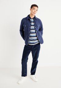 Pantalons en velours côtelé marine et une veste à deux poches assortie sur un polo rayé bleu. Des baskets blanches complètent la tenue. Design simple et décontracté.