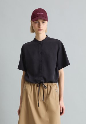 Femme portant une casquette bordeaux, une chemise noire ample à manches courtes avec des boutons, et une jupe beige avec cordon de serrage et poches zippées.