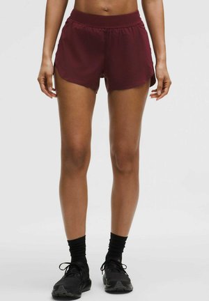 Kvinde iført mørkerøde træningsshorts, sorte sokker og sorte løbesko, stående mod en ensartet lys baggrund.