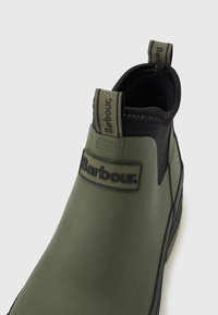 Zöld gumibakancs fekete rugalmas oldalakkal és húzófülekkel, amelyeken a "Barbour" márkajelzés látható elöl és a saroknál.