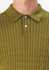 Polo de punto en verde oliva con textura acanalada, que presenta un cuello y una tapeta de tres botones. Detalle intrincado de patrón de punto en cable.