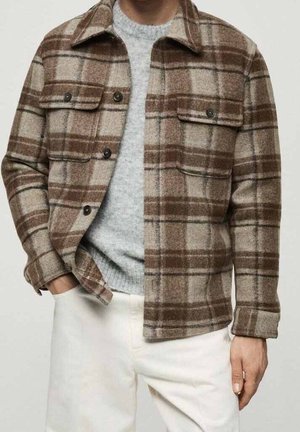 Mann trägt eine braun-beige karierte Knopfjacke über einem hellgrauen Pullover, kombiniert mit offwhitefarbenen Hosen, Hand in der Tasche.