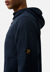 Navy blue jacket with a hood, made from lightweight fabric. Features a front pocket and a black square patch with gold logo on the sleeve.