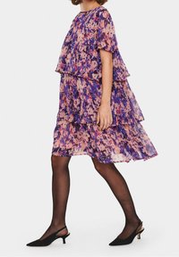 Abito a balze con stampa floreale in tessuto trasparente viola e rosa, dalla vestibilità ampia, abbinato a décolleté slingback nere e calze trasparenti.