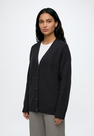 Femme aux longs cheveux foncés portant un cardigan noir boutonné par-dessus une chemise blanche et un pantalon gris, debout devant un fond clair uni.