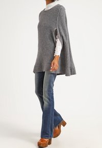 Grå stickad poncho med sidofickor, ribbad nederkant, över en vit blus, kombinerad med blå utsvängda jeans och bruna träklackar.