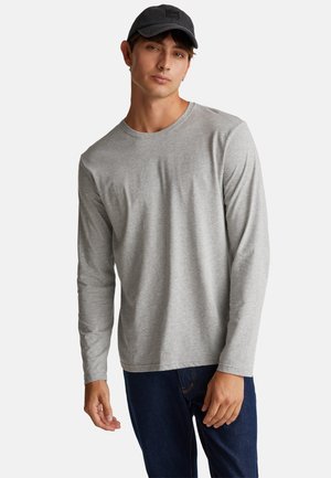 Grijze langemouwen T-shirt van een zachte stof met een ronde hals. Model die het draagt, poseert casual. Donkere baseballpet op het hoofd, spijkerbroek zichtbaar.