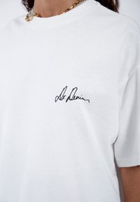 Camiseta de algodón blanca con mangas cortas que presenta un logo negro en estilo manuscrito en el lado izquierdo del pecho. Textura suave.