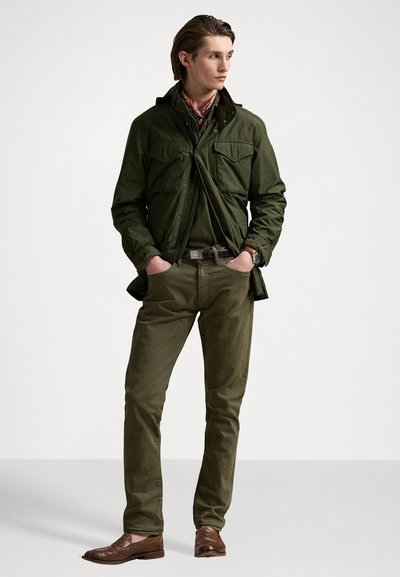 Grüne Zip-Jacke mit Brusttaschen, kombiniert mit olivfarbenen Hosen und braunen Lederschuhen. Das Outfit zeichnet sich durch einen schichten Look und Strukturvariationen aus.