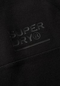 Sudadera negra con un logo "SUPER DRY®" texturizado y en relieve. Presenta una tela suave, costuras reforzadas y un diseño minimalista.
