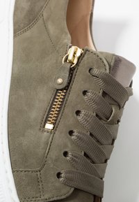 Zielona oliwkowa zamszowa sneakers z złotym zamkiem błyskawicznym, płaskimi sznurowadłami i teksturowanym akcentem z tkaniny przy pięcie. Biała gumowa podeszwa.