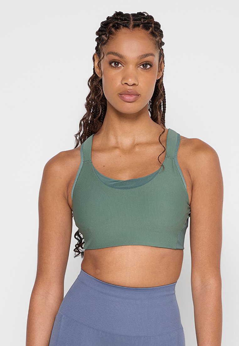 Femme avec des cheveux tressés portant un soutien-gorge de sport vert et un legging bleu taille haute, se tenant contre un fond clair.