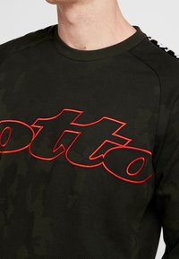 Uomo che indossa un felpa mimetica verde scuro con il testo stilizzato "lotto" in rosso sul petto e nastro di marca bianco e nero sulle spalle.