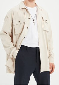 Beige overhemd met knopen, voorzien van twee borstzakken, gemaakt van lichtgewicht stof en in een relaxte pasvorm, gecombineerd met een wit T-shirt en donkere broek.