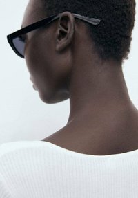 Black sunglasses with angular design, worn on a person with short, textured hair; a white ribbed top is partially visible from behind.
