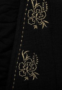 Črn material s zapletenim zlatim cvetličnim vezenjem ob robu, z teksturiranim šivanjem, ki poudarja dizajn in dodaja vizualno zanimivost.