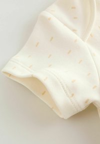 Világos krémszínű szövet apró bézs pöttyökkel, puha textúrával és hajtott mandzsettás kialakítással. Látható varrások növelik a tartósságot.