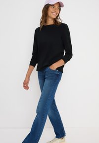 Zwart langemouw top met een relaxte fit, gecombineerd met blauwe spijkerbroek en beige sneakers. Een lichtroze pet maakt de outfit compleet.