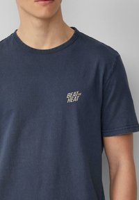 Navyblauwe katoenen t-shirt met een ronde hals. Bevat het logo "BEAT the HEAT" in lichtbeige op de linkerkant van de borst. Korte mouwen en een zachte textuur.