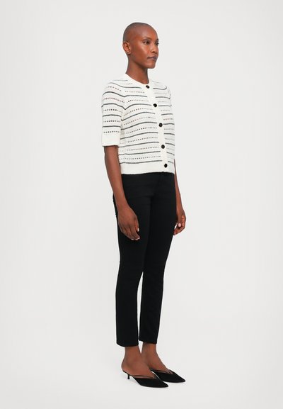 Witte gebreide cardigan met korte mouwen, voorzien van horizontale strepen en knoopsluitingen, gecombineerd met zwarte slim-fit broek en zwarte hakken.