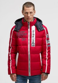 Red puffer jacket with a navy hood. Features white stripes, text graphics, zip pockets, and a quilted texture. Notable branding on the front.
