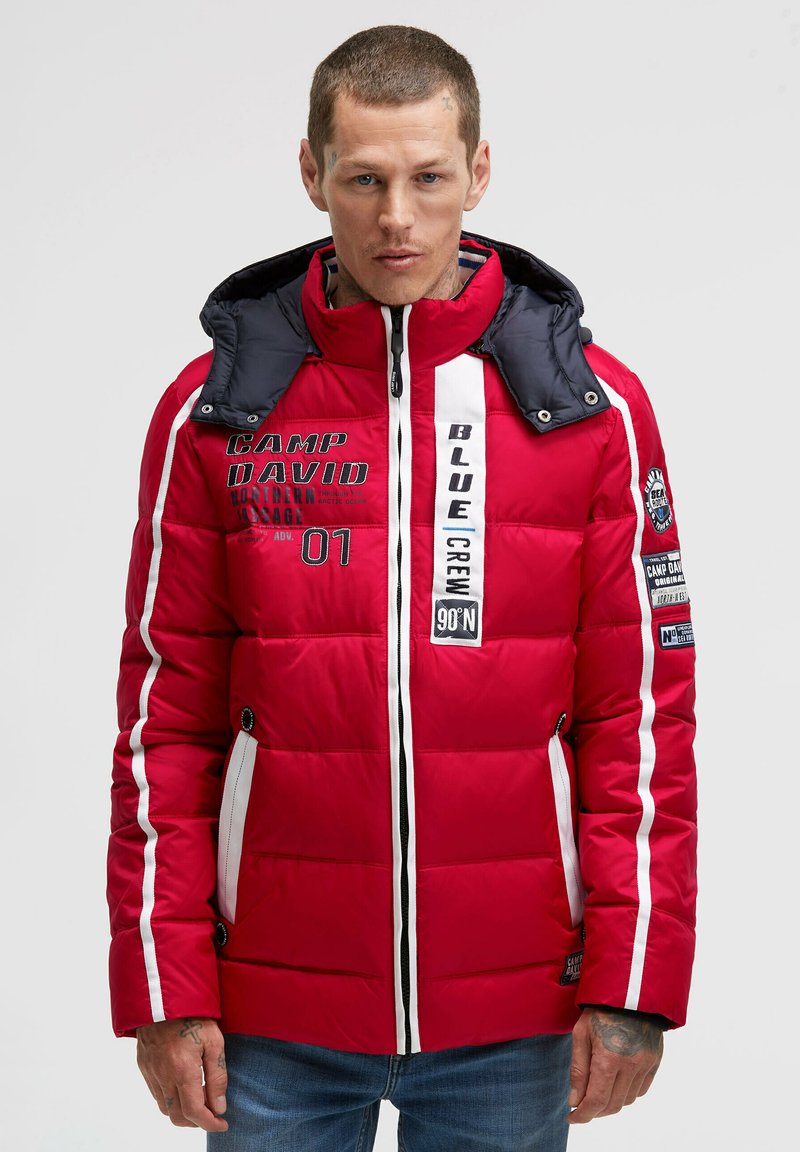 Red puffer jacket with a navy hood. Features white stripes, text graphics, zip pockets, and a quilted texture. Notable branding on the front.