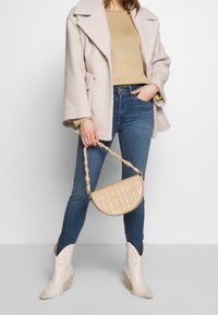 Lichtgekleurde, gestructureerde oversized jas over een beige sweater, gecombineerd met een passende spijkerbroek en kniehoog crème cowboylaarzen. Draagt een beige handtas met krokodillenpatroon.