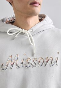 Világosszürke kapucnis pulóver, hímzett "Missoni" logóval, amely színes zig-zag mintákkal díszített. Fehér zsinórral, fém végekkel.