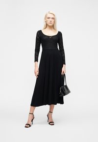 Γυναίκα με μαύρο δαντελωτό φόρεμα με μακριά μανίκια, κρατώντας μαύρη τσάντα χειρός και φορώντας μαύρα πέδιλα με ψηλό τακούνι, στέκεται σε λευκό φόντο.