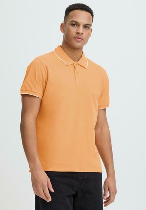 Mann trägt ein oranges kurzärmeliges Poloshirt mit weißer Einfassung und schwarze Hose, steht vor einem schlichten hellen Hintergrund.