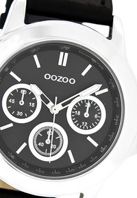 Montre analogique noire avec boîtier argenté, trois sous-cadrans et marquages blancs. Elle possède un bracelet noir lisse et une échelle tachymétrique autour de la lunette.